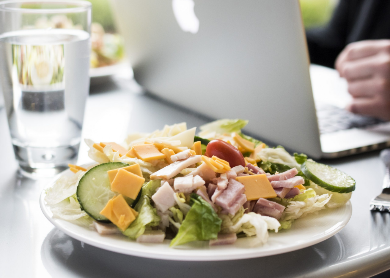 A plate of salad with lettuce, cucumber, tomato, cheese, and bacon sits next to a glass of water and an open laptop in a bright dining setting—ideal for busy employers or Continental Services’ meal breaks.