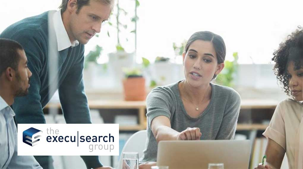 Four professionals are having a discussion around a table. One woman is pointing at a laptop screen while others listen, highlighting the spirit of partnership. The Execu|Search Group logo is displayed in the lower left corner.