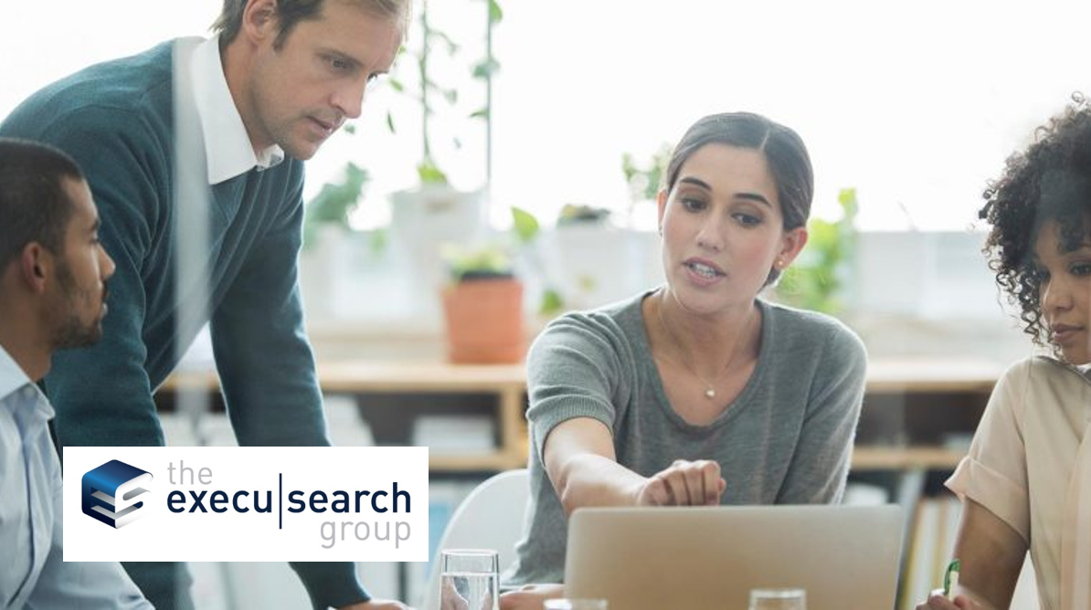 Four people in business attire discuss ideas around a table with a laptop. One woman gestures while speaking. The logo “the execu|search group” appears in the bottom left, highlighting connections to Covalent Health.