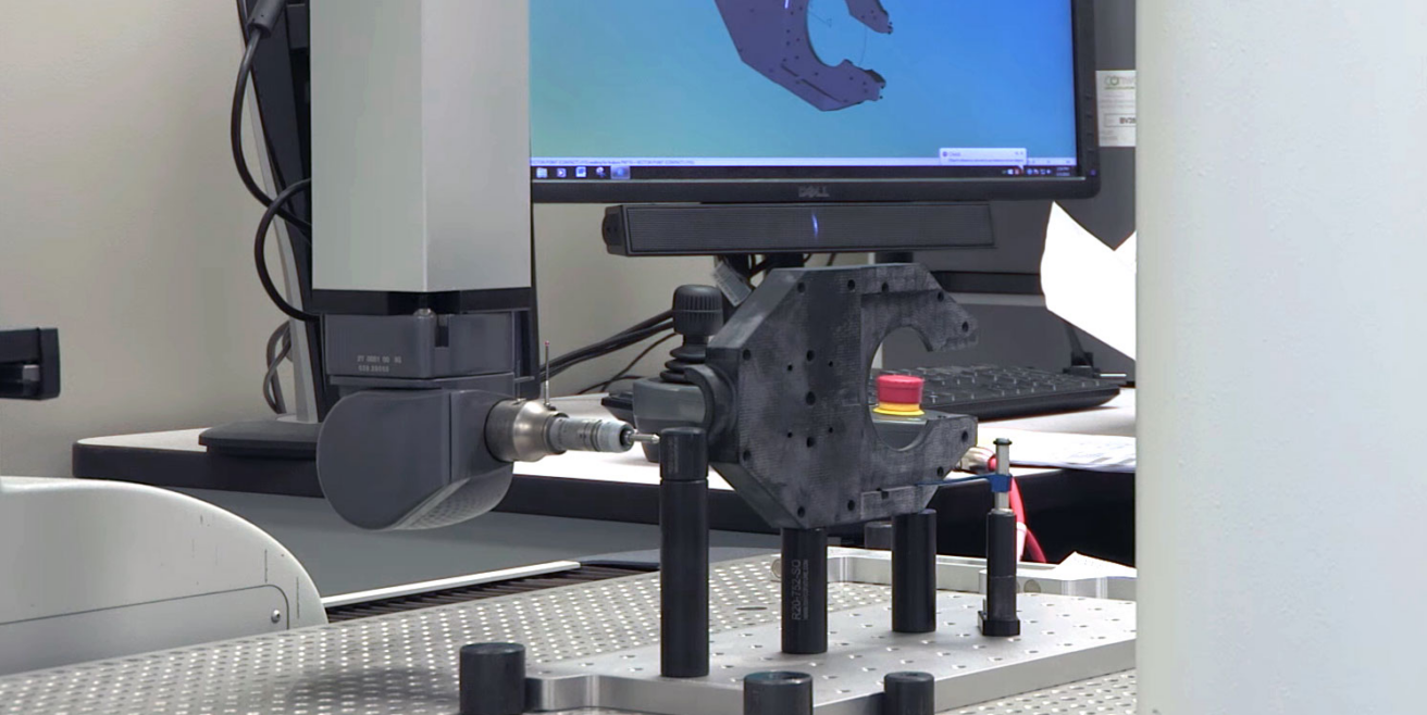 A coordinate measuring machine (CMM) probe inspects a black mechanical part on a metal table, with a computer monitor in the background displaying a 3D model—showcasing EPTAM Plastics’ commitment to quality and precision as an ongoing investment.