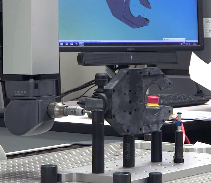 A coordinate measuring machine (CMM) probe inspects a black mechanical part on a metal table, with a computer monitor in the background displaying a 3D model—showcasing EPTAM Plastics’ commitment to quality and precision as an ongoing investment.