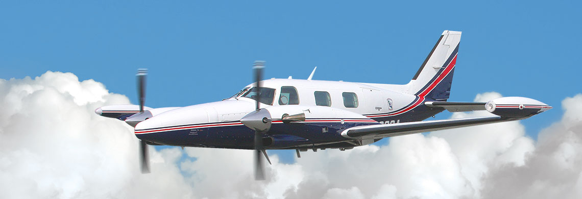A small twin-engine propeller airplane with a white and blue body and red stripes, operated by REACH Air Medical Services, flies in front of white clouds against a bright blue sky.