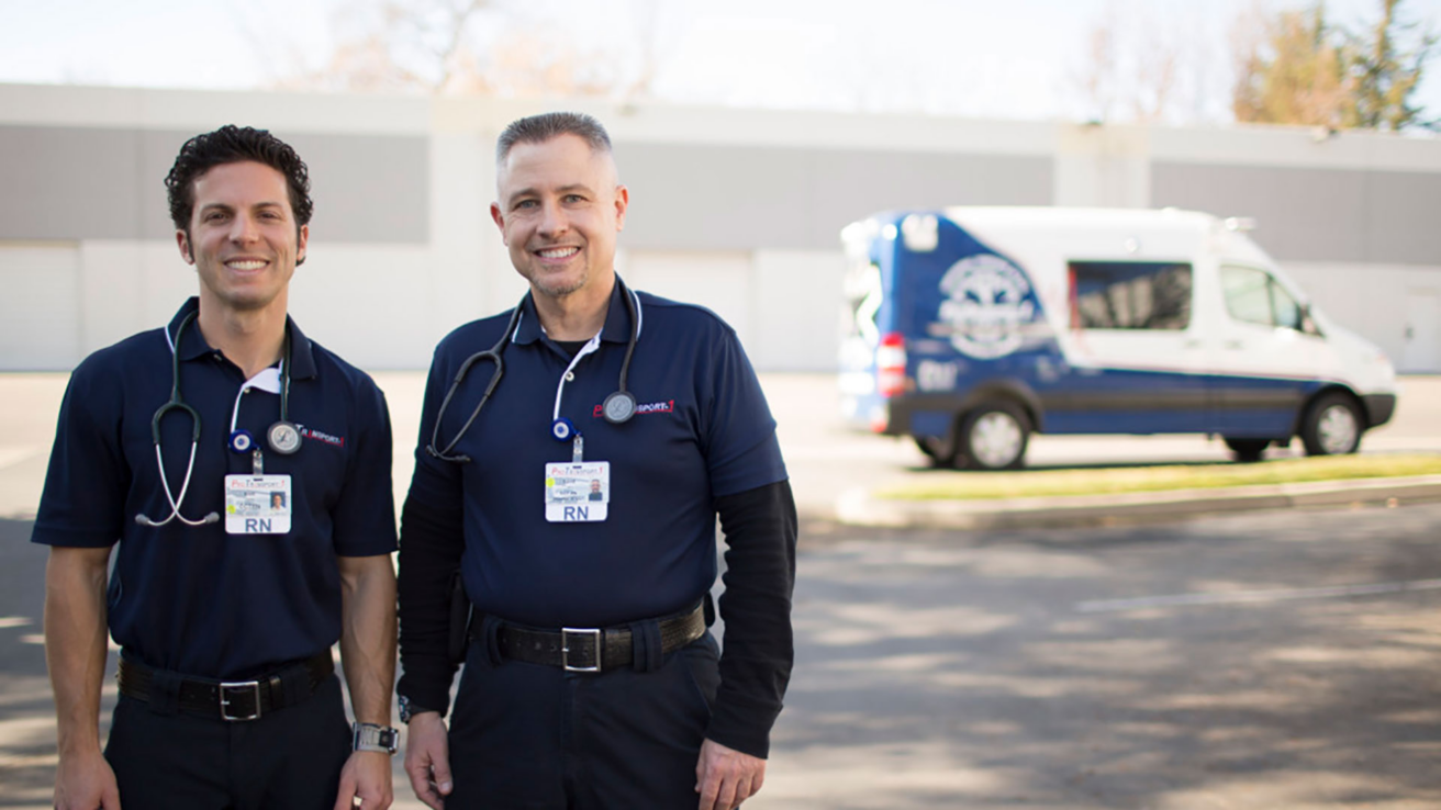 Two male nurses in navy blue uniforms and stethoscopes stand outside, smiling, with a ProTransport-1 ambulance van parked in the blurred background, reflecting a heritage of care in which the company proudly invests.