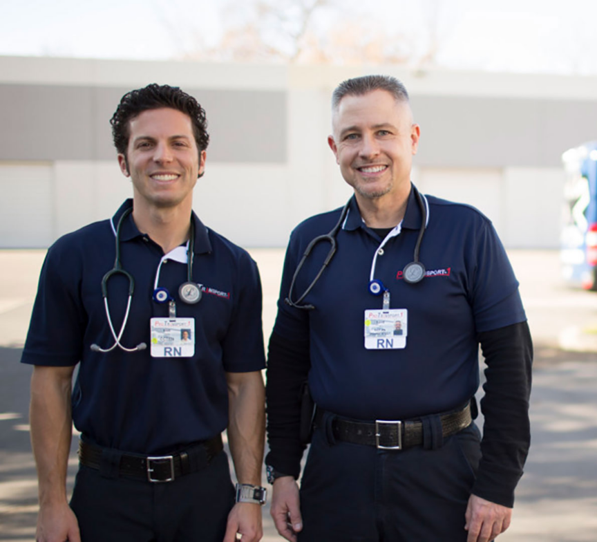 Two male nurses in navy blue uniforms and stethoscopes stand outside, smiling, with a ProTransport-1 ambulance van parked in the blurred background, reflecting a heritage of care in which the company proudly invests.