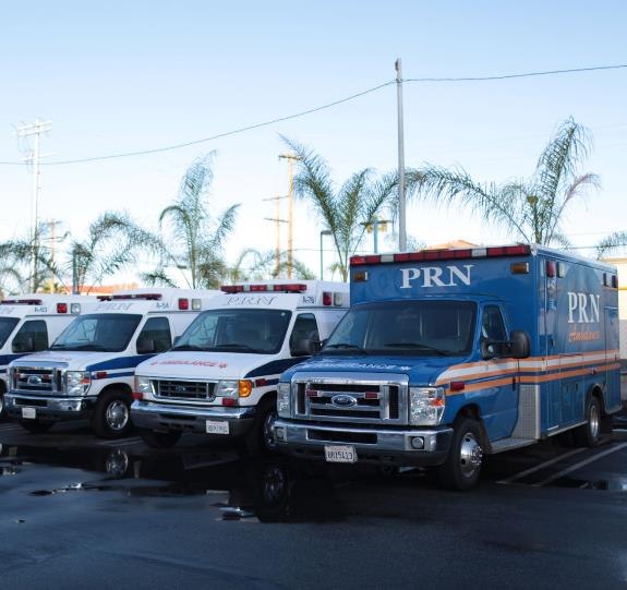 Four ambulances, including a blue PRN Ambulance and three white ones, are parked side by side in a lot with palm trees and power lines visible in the background after ProTransport-1 acquires PRN Ambulance.