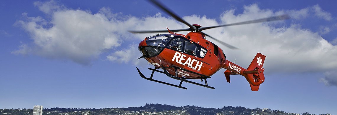 A red REACH Air Medical Services helicopter soars above a hilly landscape dotted with buildings, flying under scattered clouds on a sunny day.
