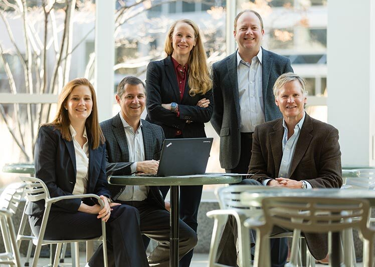 Five people in business attire, including Melissa Barry, pose and smile around a small table with a laptop in a bright, modern office with large windows—a space that reflects both professionalism and heritage.