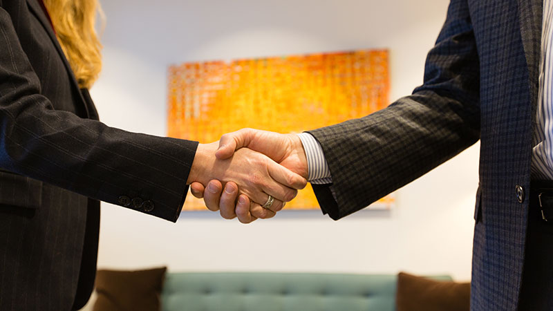 Two people in business attire shake hands in an office setting, symbolizing a successful minority investing partnership. A blurred orange and yellow painting adds a vibrant touch in the background.