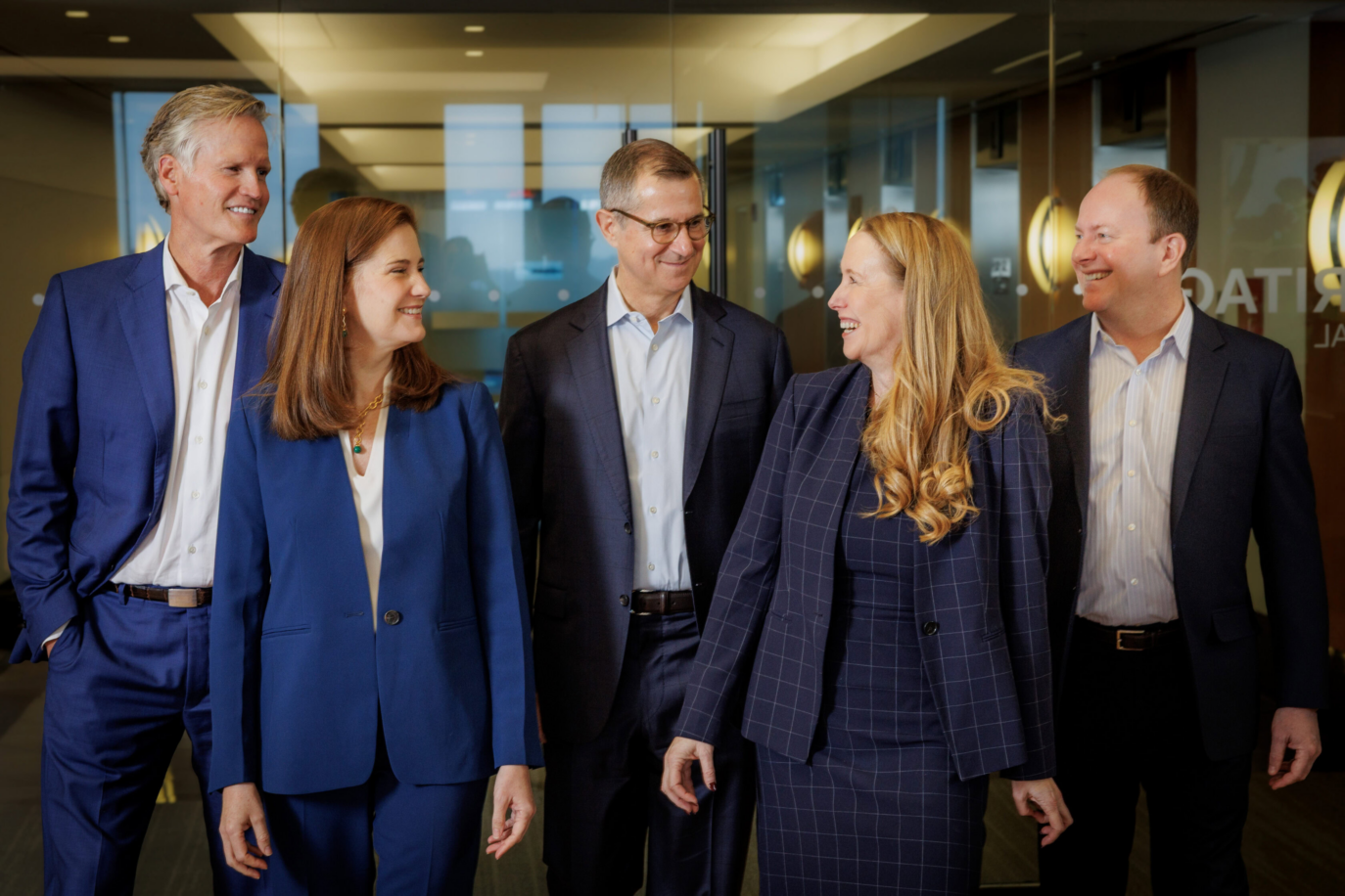 Five business professionals, three men and two women, stand together in formal attire, smiling and talking in a modern office hallway with glass walls and warm lighting, capturing a moment for the Staff Archive.