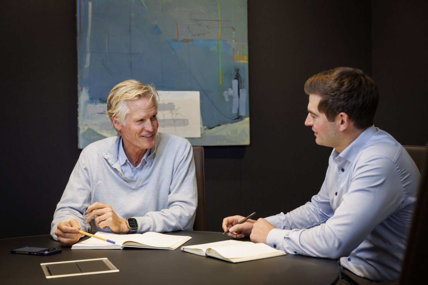 Two men sit at a round table in an office, engaged in conversation. With notebooks and pens from the Staff Archive in front of them, they discuss ideas beneath a colorful abstract painting on the dark wall behind them.