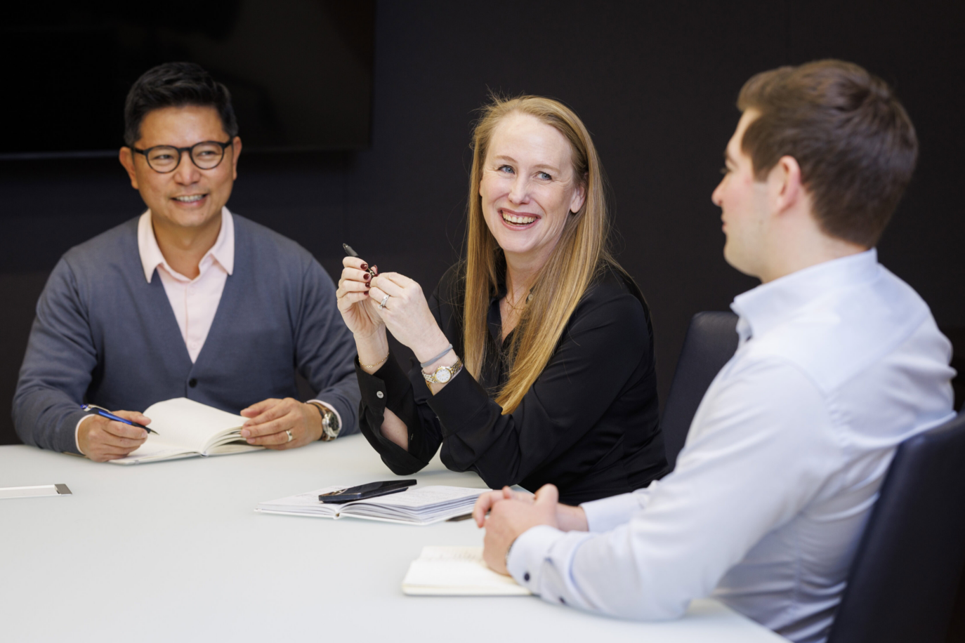 Three people sit at a conference table with notebooks, engaged in a meeting. The woman in the center is smiling and holding a pen, while the two men on either side listen attentively, creating a moment perfect for the Staff Archive.