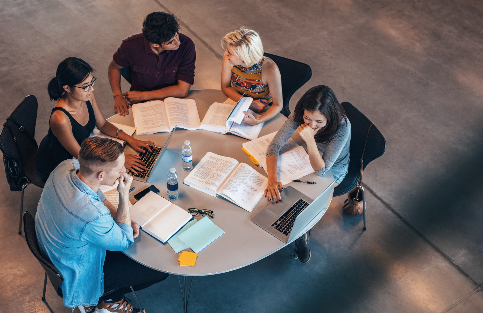 Five people sit around a round table with laptops, books, notebooks, and water bottles, engaged in discussion and studying together in a modern, open space.