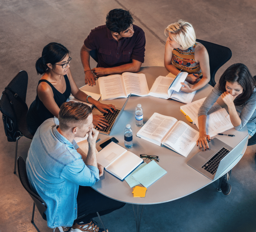Five people sit around a round table with laptops, books, notebooks, and water bottles, engaged in discussion and studying together in a modern, open space.