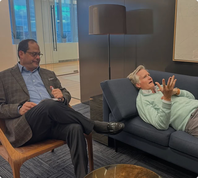 Two men are having a conversation in an office. One sits on a wooden chair, listening, while the other reclines on a dark sofa, gesturing as he speaks—like hosts recording podcasts for an archive. A tall lamp and large window are in the background.