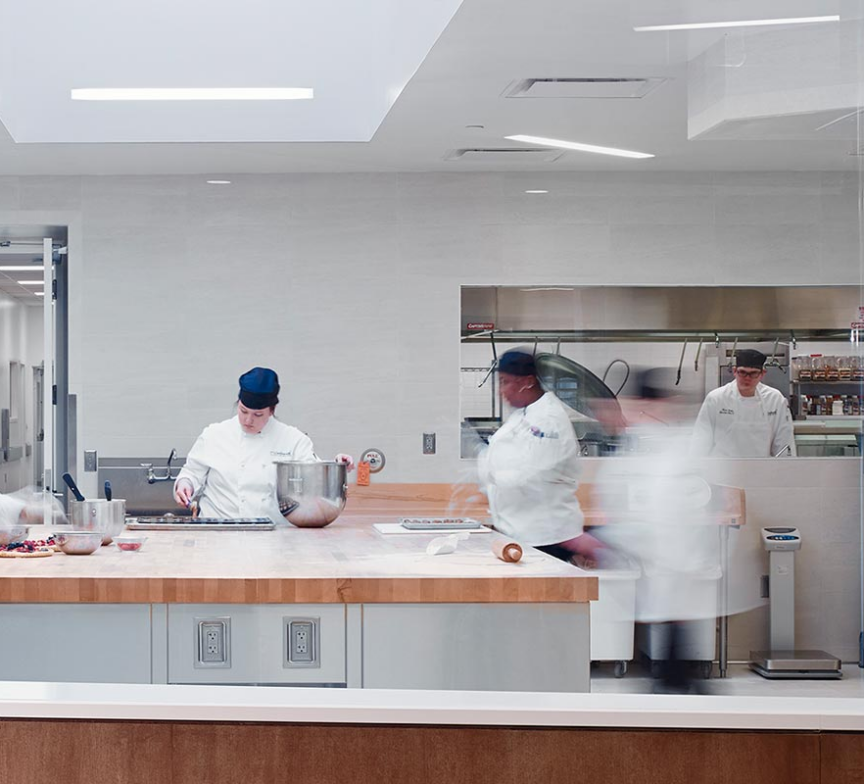 Four pastry chefs in white uniforms and dark hats work in a bright, modern bakery kitchen for Continental Services. Some mix ingredients while others handle dough at a large wooden counter, with fresh bread visible on racks in the background.