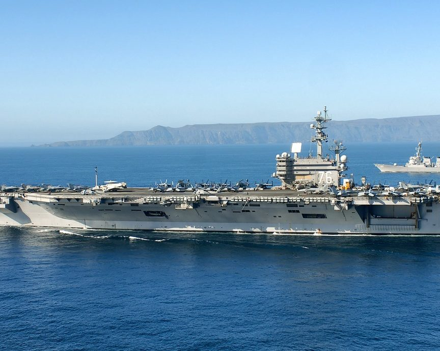 A large aircraft carrier sails on blue ocean water with numerous jets on its deck, accompanied by a smaller naval ship in the background, and an island is visible on the horizon under a clear sky.