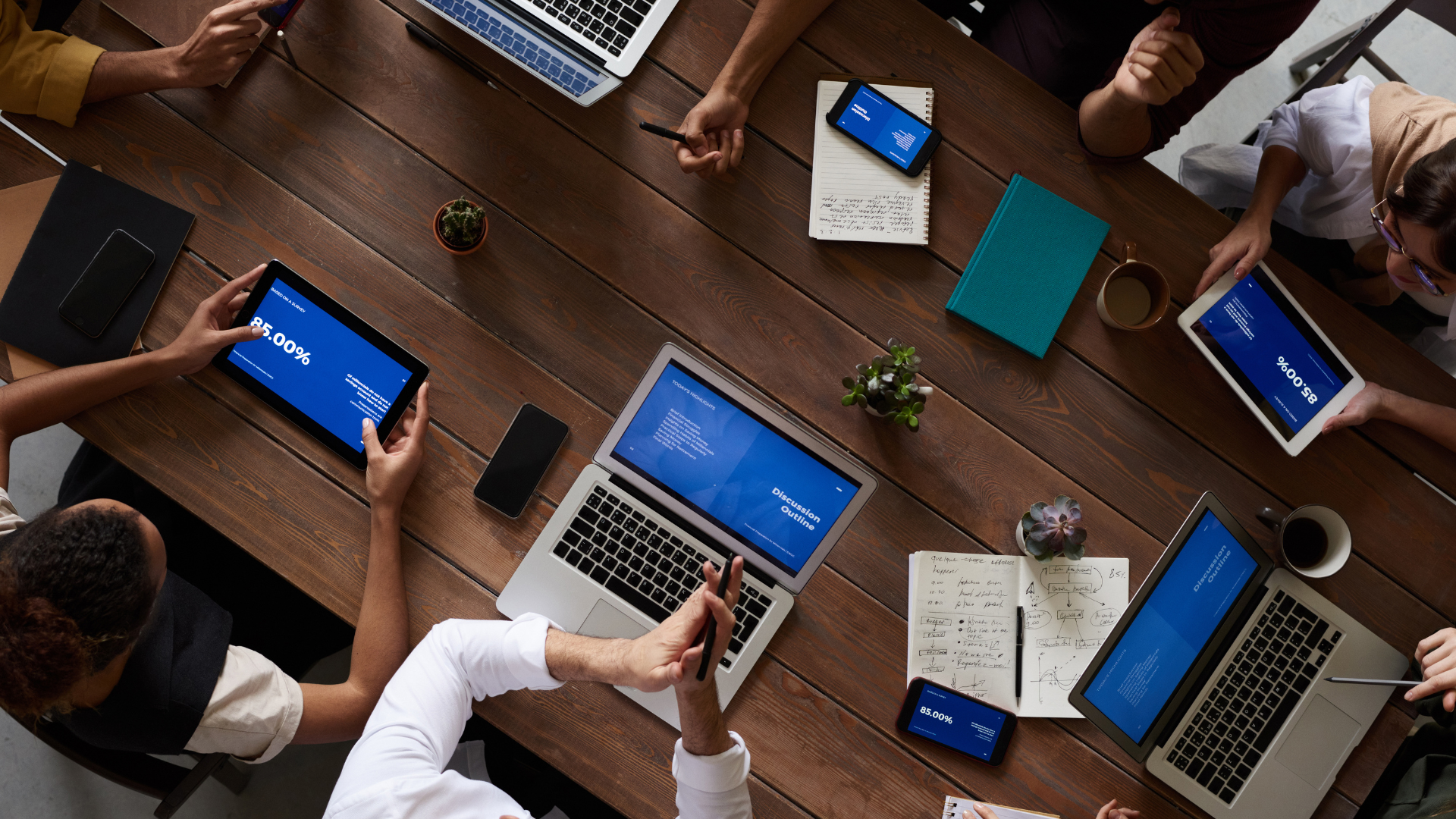 A group of people sit around a wooden table with laptops, tablets, notebooks, and phones, collaborating and discussing during a meeting. Screens display charts and text, and some hands gesture or take notes.