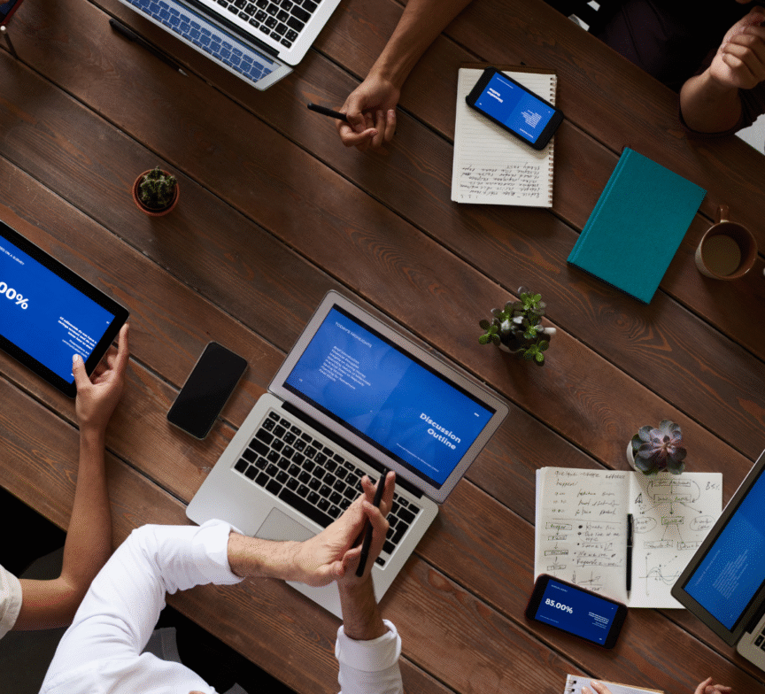 A group of people sit around a wooden table with laptops, tablets, notebooks, and phones, collaborating and discussing during a meeting. Screens display charts and text, and some hands gesture or take notes.