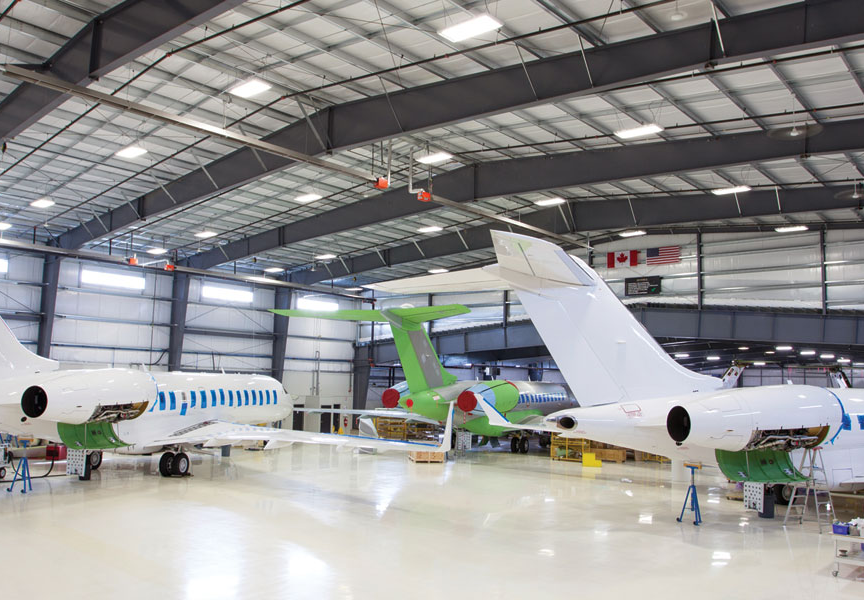 Two white private jets and one green jet, maintained by Flying Colours Corp, are parked inside a spacious, well-lit aircraft hangar with high ceilings, large windows, and visible maintenance equipment around the planes.