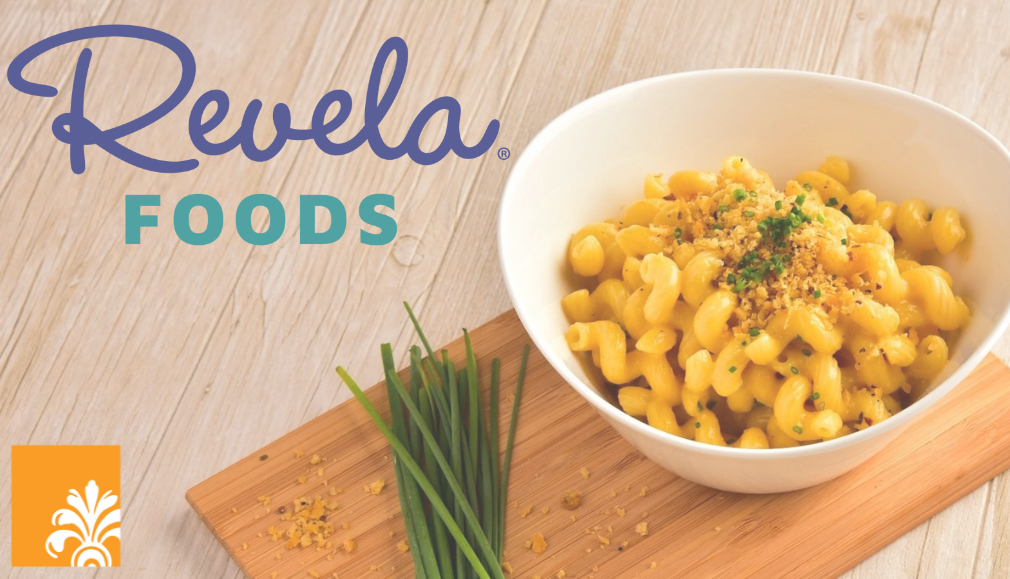 A bowl of macaroni and cheese topped with herbs sits on a wooden board with chives. The "Revela Foods" logo appears on the left, highlighting their success following an investment exit by Heritage Capital, alongside an orange square with a white floral design.