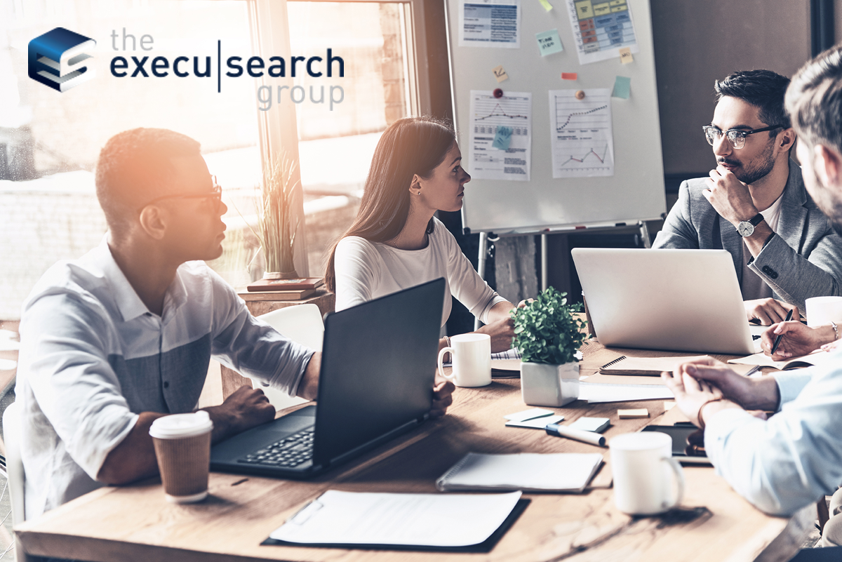 Three professionals sit at a table with laptops, coffee, and documents, discussing TechLink’s recent acquisition. A woman faces two men in an office with charts on a whiteboard. The Execu|Search Group logo appears in the top left corner.