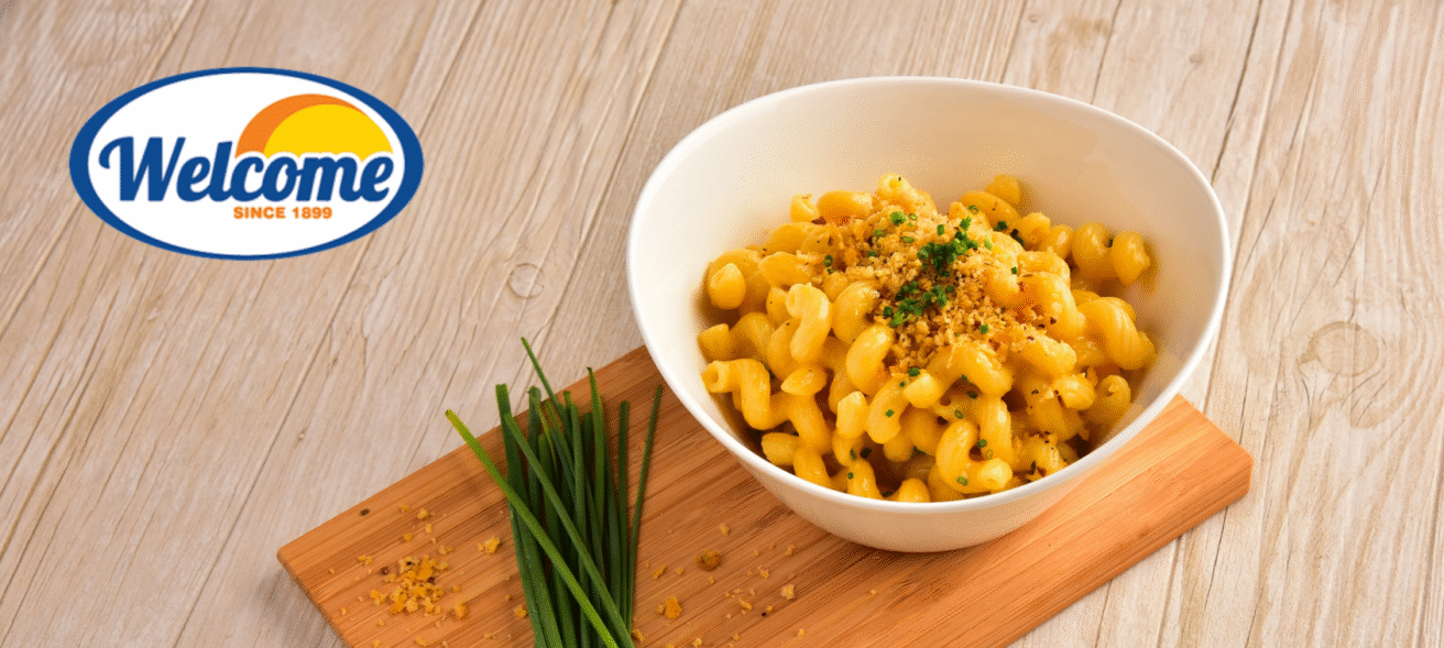 A bowl of macaroni and cheese garnished with chopped herbs sits on a wooden board next to fresh chives, with a "Welcome Dairy" logo in the top left corner on a light wood table.
