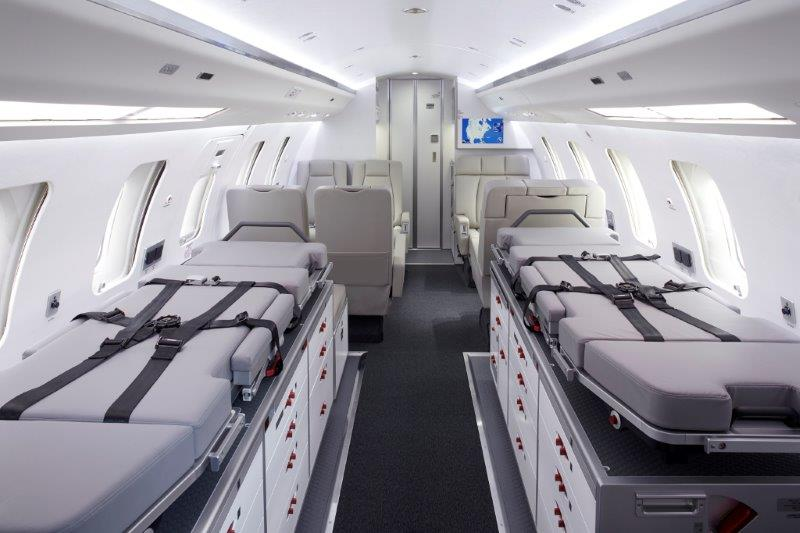 The image shows the interior of a Bombardier Challenger 650 medevac jet with two patient stretchers, medical equipment storage units, and several passenger seats in a clean, well-lit cabin by Flying Colours Corp.