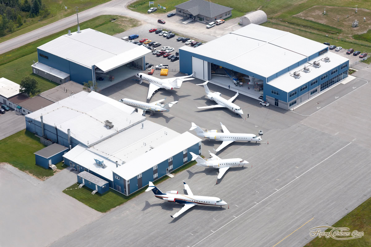 Aerial view of Peterborough airport facility with several small private jets parked on the tarmac near blue and white Flying Colours hangars, cars parked nearby, and grassy areas surrounding the buildings.