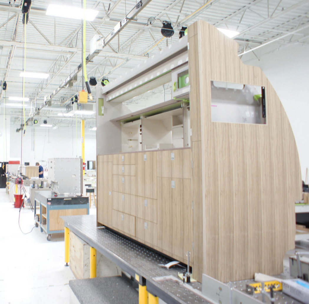 A large wooden structure, possibly a custom cabinet or interior module, stands in the middle of Flying Colours Corp's bright, modern St. Louis facility, with exposed ceilings and active production workstations in the background.