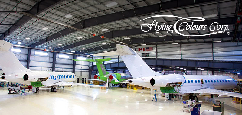 Interior of an aircraft hangar with three private jets being serviced. The planes, painted white with blue and green accents, showcase the signature style of Flying Colours Corp. The logo is displayed in the upper right corner.