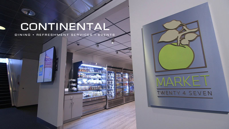 Modern indoor market with refrigerated food displays, a wall-mounted sign reading "Market Twenty 4 Seven" with a green apple graphic, and "Continental Services · Refreshment Services · Events" text overlay—a Satellite Vending acquisition.