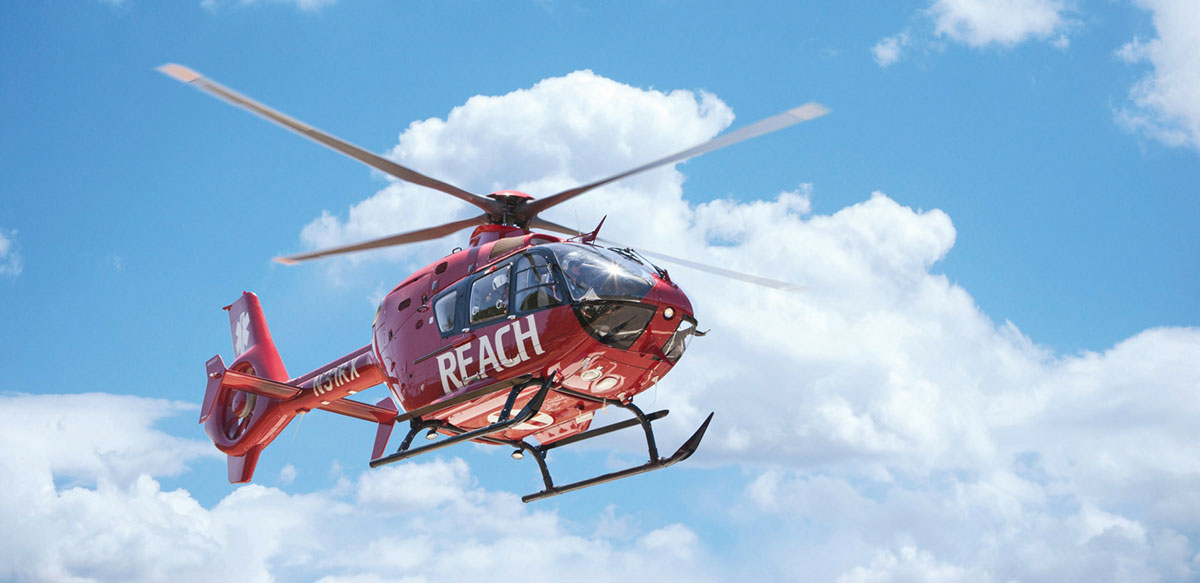 A red medical helicopter with the words "REACH Air Medical Services" on its side is flying in a blue sky with scattered clouds.