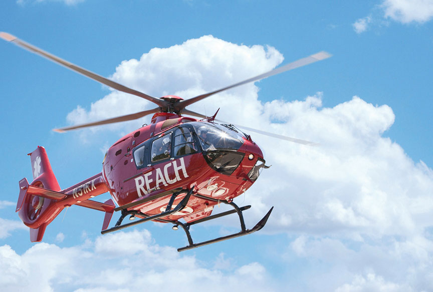 A red medical helicopter with the words "REACH Air Medical Services" on its side is flying in a blue sky with scattered clouds.