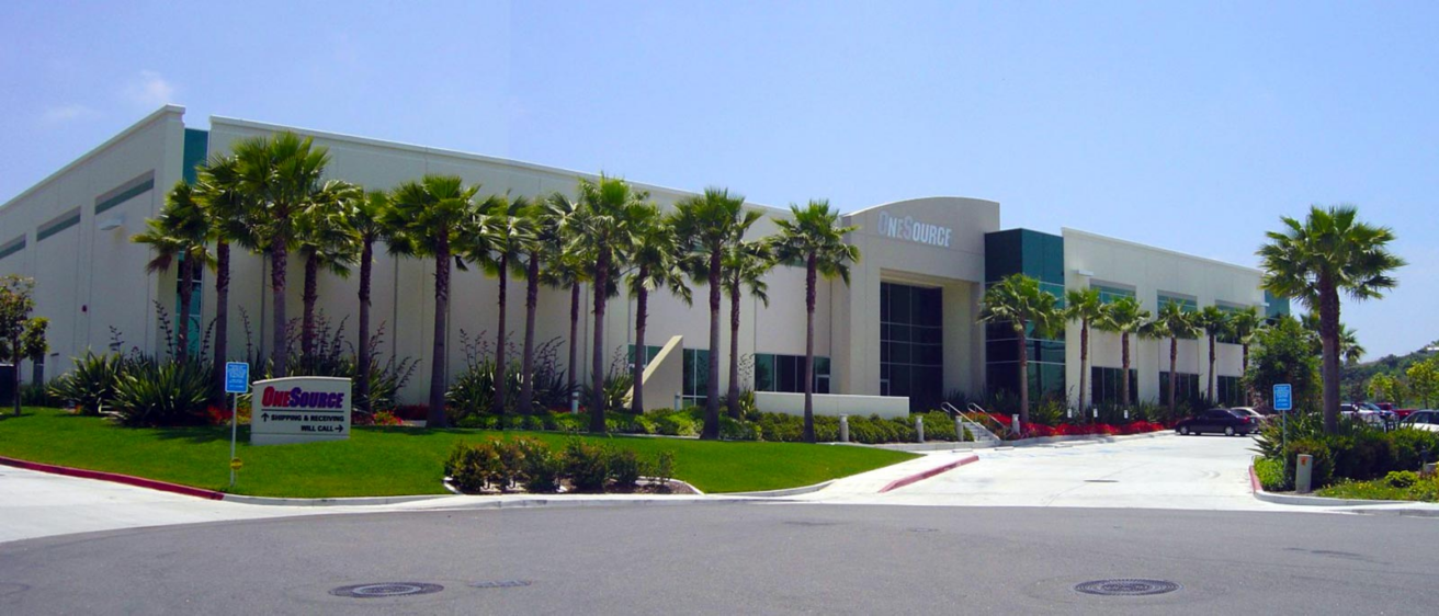 A modern, white commercial building with tall windows and palm trees lining the sidewalk houses OneSource Distributors, a Sonepar USA company. The area is clean and well-maintained under a clear blue sky.