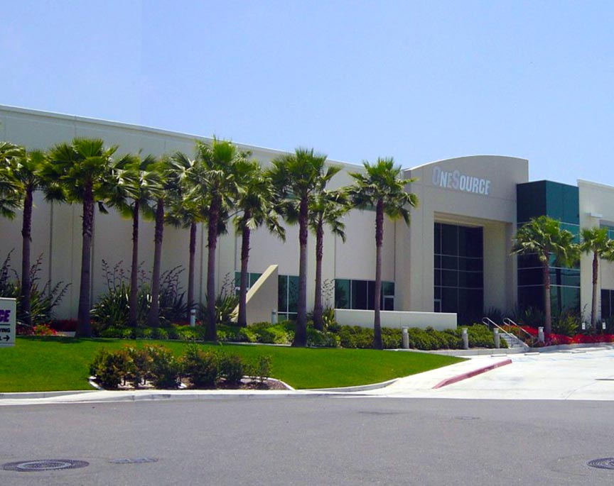 A modern, white commercial building with tall windows and palm trees lining the sidewalk houses OneSource Distributors, a Sonepar USA company. The area is clean and well-maintained under a clear blue sky.