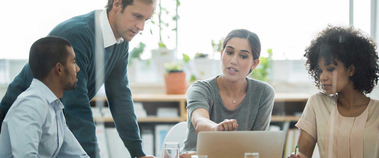 Four people sit around a table in an office, engaged in discussion. One woman from Execu I Search Group gestures toward a laptop screen while the others listen attentively. The collaborative atmosphere reflects the team's commitment to investing in shared success.
