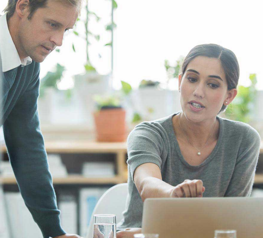 Four people sit around a table in an office, engaged in discussion. One woman from Execu I Search Group gestures toward a laptop screen while the others listen attentively. The collaborative atmosphere reflects the team's commitment to investing in shared success.