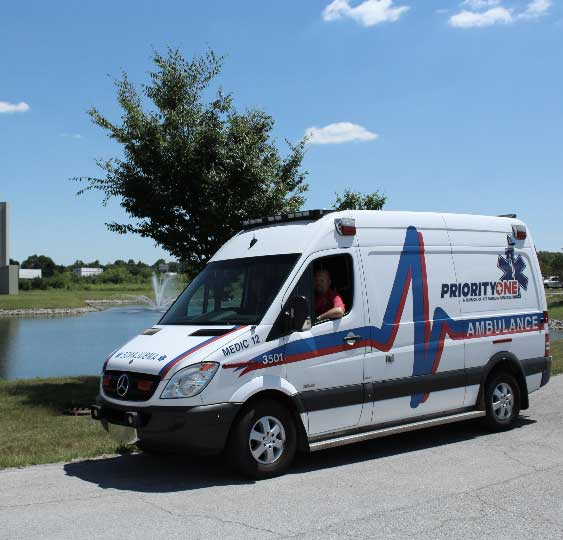 A Priority One ambulance, now part of ProTransport-1 after a recent acquisition, is parked by a pond on a sunny day. A tree and fountain add to the scene while a person sits in the driver’s seat.