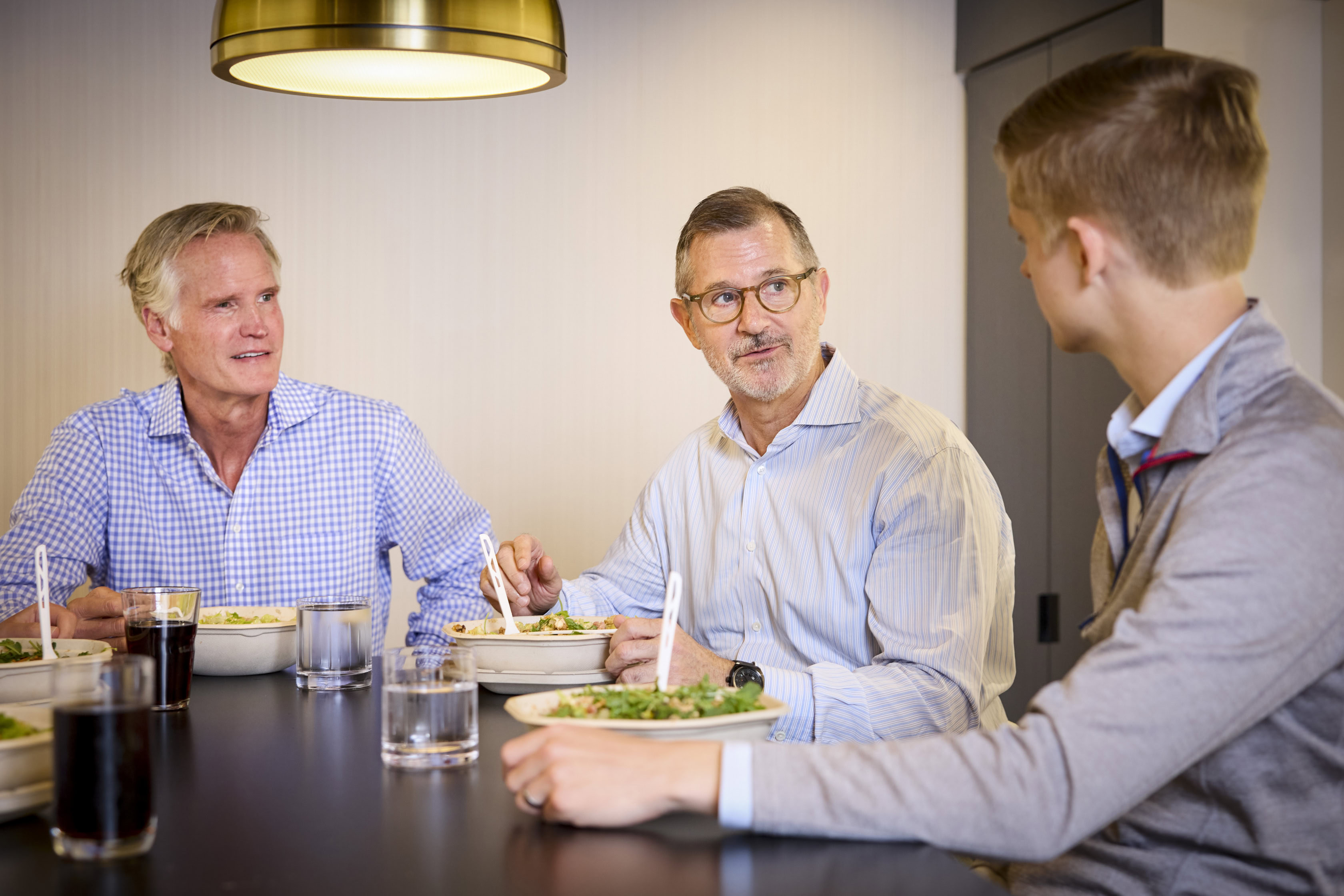 Three men sit at a dining table, eating salads and talking. Two have gray hair and glasses, while the third is a young man with short blond hair. Glasses of water and soda rest on the table under a hanging light in this Staff Archive scene.