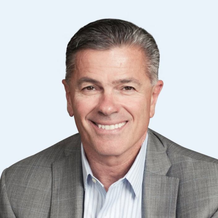 A middle-aged man with short gray hair, wearing a gray checked suit jacket over a light blue collared shirt, smiles at the camera against a plain light background—reflecting the professional spirit of Cascadia Capital.