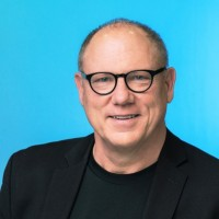 A middle-aged man with short light hair and glasses smiles at the camera. He wears a black blazer over a black shirt and is posed in front of a bright blue background, representing By the People Technologies with a professional, approachable presence.