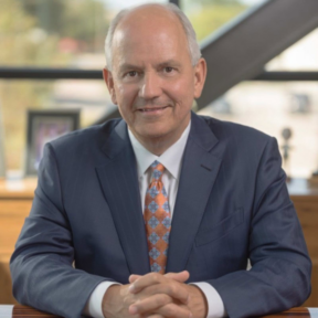 A man in a blue suit and patterned orange tie sits at a desk with his hands clasped, smiling at the camera. The background shows large windows and blurred office decor.