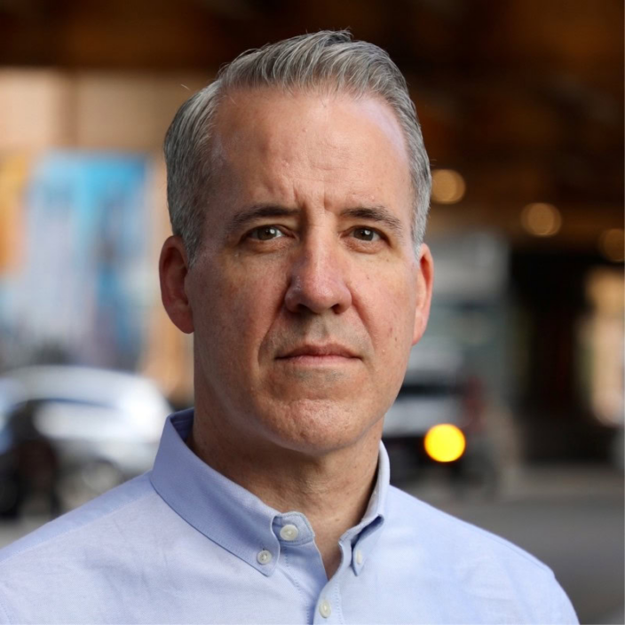 A middle-aged man with short gray hair wearing a light blue button-up shirt stands outdoors, looking directly at the camera with a serious expression. The blurred background includes cars and urban scenery.