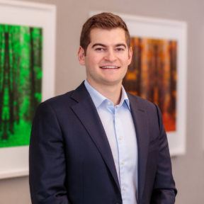 A man in a dark suit and light blue shirt smiles while standing indoors. Behind him, two framed photographs of forest scenes—one green and one orange—hang on the wall.