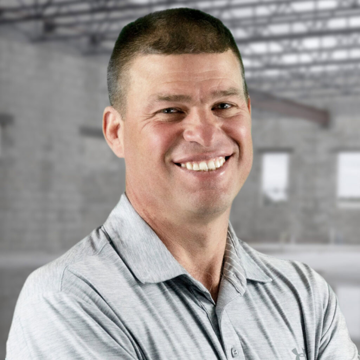 A smiling man with short brown hair wearing a light gray collared shirt stands with arms crossed in front of a blurred, unfinished building interior.