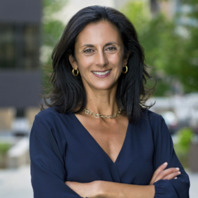 A woman with long dark hair stands outdoors, smiling with her arms crossed. She is wearing a navy blouse, gold hoop earrings, a gold necklace, and a bracelet, with a blurred cityscape background.