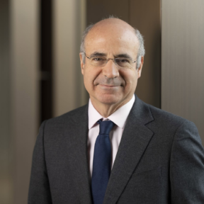 A middle-aged man with glasses, wearing a dark suit, white shirt, and blue tie, stands in front of a blurred office background, smiling softly at the camera.