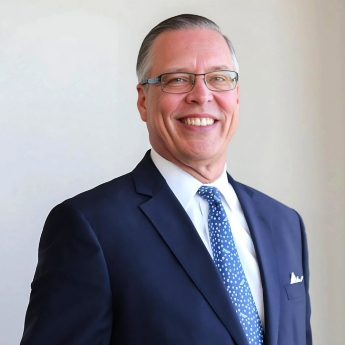 A smiling man wearing glasses, a dark blue suit, a white shirt, and a blue patterned tie stands against a plain light-colored background.