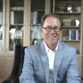 A smiling man with glasses and a light blue plaid suit jacket sits in a chair in front of bookshelves with glass doors.