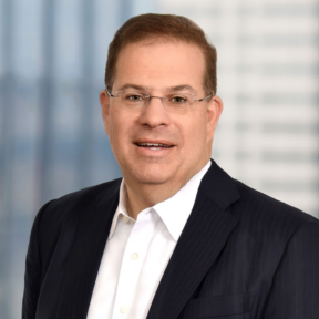 A man wearing glasses, a dark pinstripe suit, and a white dress shirt, smiling at the camera with a blurred office background.